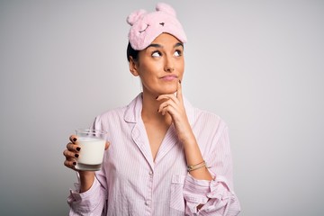 Young beautiful woman wearing pajama and sleep mask drinking glass of healthy milk serious face thinking about question, very confused idea