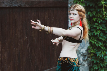 beautiful woman with long red hair in green tribal dance costume with outstretched arms looking at camera and standing near wooden gate