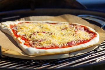 A closeup portrait of a pizza being baked on a pizza stone on a barbecue grill. The pizza is topped with cheese, tomato sauce and other toppings.