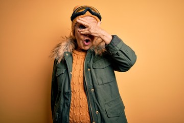 Senior beautiful grey-haired skier woman wearing snow sportswear and ski goggles peeking in shock covering face and eyes with hand, looking through fingers with embarrassed expression.