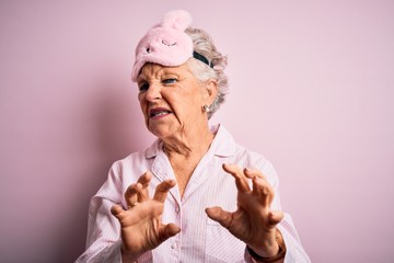 Senior beautiful woman wearing sleep mask and pajama over isolated pink background disgusted expression, displeased and fearful doing disgust face because aversion reaction. With hands raised
