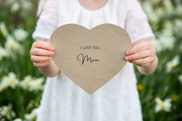 I love you mum written on a heart-shaped card held by a girl on flower and grass background