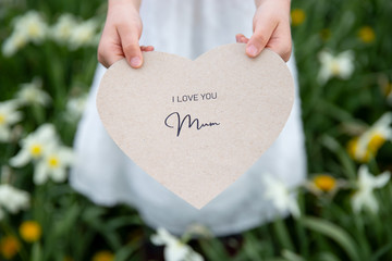 I love you mum written on a heart-shaped paper card by a girl on grass background