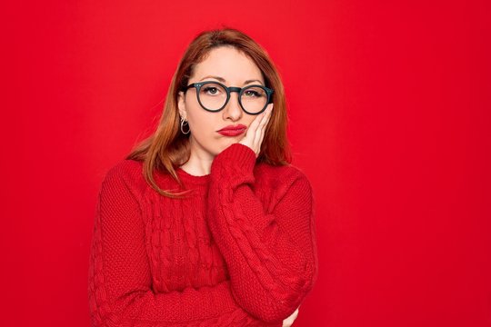 Young beautiful redhead woman wearing casual sweater and glasses over red background thinking looking tired and bored with depression problems with crossed arms.
