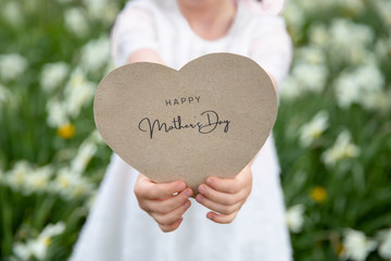 Happy Mother's Day written on a heart-shaped card held by a girl on flower and grass background