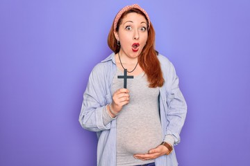 Young beautiful redhead pregnant woman expecting baby holding christian cross scared in shock with a surprise face, afraid and excited with fear expression