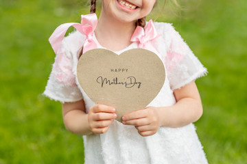 Happy Mother's Day written on a heart-shaped card held by a girl on grass background