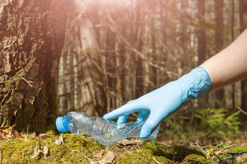 An empty crumpled plastic bottle is lying on the ground in the forest. Hand in a blue rubber glove. The concept of garbage, forest cleaning, negative impact on nature, ecology, environment, cleanup.