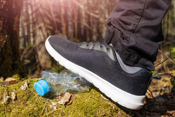 A leg in a black sneaker steps on an empty crumpled plastic bottle from the ground in the forest. The concept of garbage, the negative impact of human on nature, ecology, environment.
