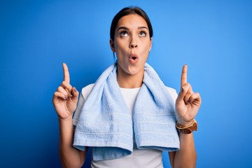 Young beautiful brunette sportswoman wearing towel doing exercise over blue background amazed and surprised looking up and pointing with fingers and raised arms.