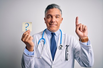 Middle age handsome grey-haired doctor man holding paper with question mark surprised with an idea or question pointing finger with happy face, number one