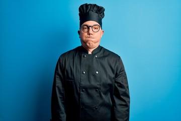 Middle age handsome grey-haired chef man wearing cooker uniform and hat puffing cheeks with funny face. Mouth inflated with air, crazy expression.