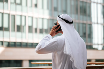 Emirati guy wearing traditional kandura in urban city emirates lifestyle talking to smart phone. Overlooking building from the balcony. Photography concept for Arab magazine.