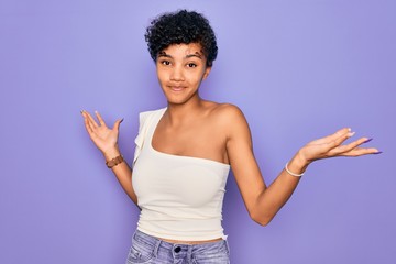 Young beautiful african american afro woman wearing casual t-shirt over purple background clueless and confused expression with arms and hands raised. Doubt concept.