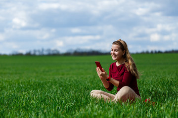 Young beautiful woman student with a phone in her hands sitting on the grass. Girl takes selfies and takes selfie pictures. She smiles and enjoys a warm day. Concept photo on smartphone