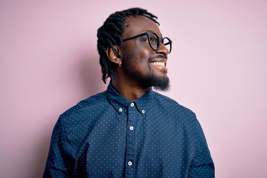 Young handsome african american man wearing casual shirt and glasses over pink background looking away to side with smile on face, natural expression. Laughing confident.