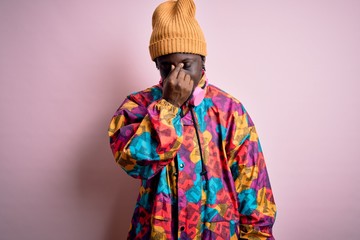 Young handsome african american man wearing colorful coat and cap over pink background tired rubbing nose and eyes feeling fatigue and headache. Stress and frustration concept.