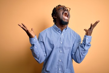 Young handsome african american man wearing shirt and glasses over yellow background celebrating mad and crazy for success with arms raised and closed eyes screaming excited. Winner concept