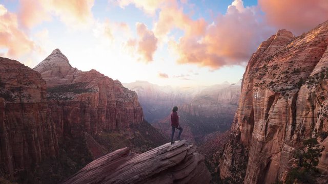 Adventurous Woman at the edge of a cliff is looking at a beautiful landscape view in the Canyon during a vibrant sunset. Taken in Zion National Park, Utah, United States. Parallax Panorama