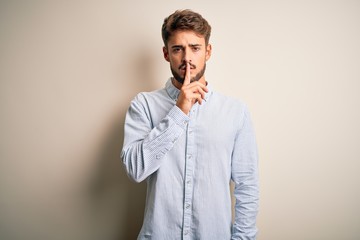 Young handsome man with beard wearing striped shirt standing over white background asking to be quiet with finger on lips. Silence and secret concept.