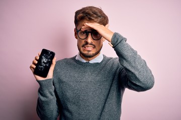 Young man with beard wearing glasses holding broken and craked smartphone stressed with hand on...