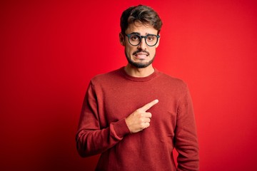 Young handsome man with beard wearing glasses and sweater standing over red background Pointing...