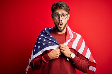 Young man wearing glasses and United States of America flag over isolated red background scared in shock with a surprise face, afraid and excited with fear expression