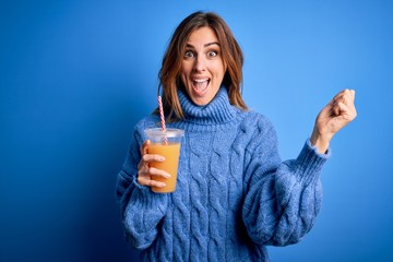 Young beautiful brunette woman drinking healthy orange juice over blue background screaming proud and celebrating victory and success very excited, cheering emotion