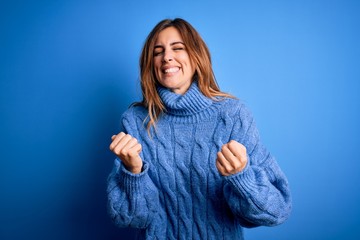 Young beautiful brunette woman wearing casual turtleneck sweater over blue background very happy and excited doing winner gesture with arms raised, smiling and screaming for success. Celebration