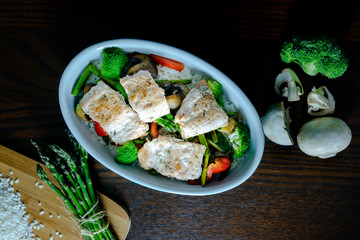 Fuente con salmón salvaje, arroz, esparragos trigueros, brócoli, pimiento rojo y champiñones sobre mesa marrón. Dark photography. Cenital.