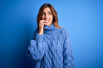 Young beautiful brunette woman wearing casual turtleneck sweater over blue background looking stressed and nervous with hands on mouth biting nails. Anxiety problem.