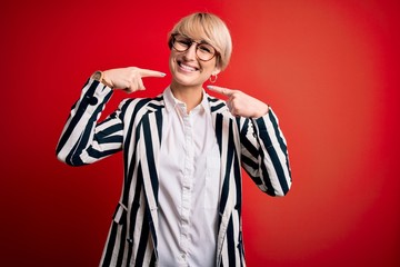 Blonde business woman with short hair wearing glasses and striped jacket over red background smiling cheerful showing and pointing with fingers teeth and mouth. Dental health concept.