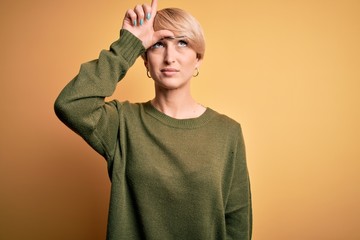 Young blonde woman with modern short hair wearing casual sweater over yellow background making fun of people with fingers on forehead doing loser gesture mocking and insulting.