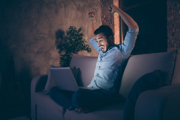 Profile side view of his he nice attractive lucky cheerful cheery glad brunet guy sitting on divan using laptop rejoicing having fun at modern loft industrial style interior dark room apartment flat
