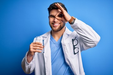 Young blond doctor man with beard and blue eyes wearing coat drinking glass of water with happy face smiling doing ok sign with hand on eye looking through fingers