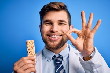Young blond doctor man with beard and blue eyes wearing coat eating granola bar doing ok sign with fingers, excellent symbol