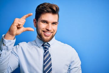 Young blond businessman with beard and blue eyes wearing elegant shirt and tie standing smiling and confident gesturing with hand doing small size sign with fingers looking and the camera. Measure