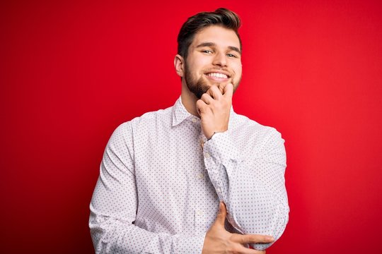 Young blond businessman with beard and blue eyes wearing elegant shirt over red background looking confident at the camera with smile with crossed arms and hand raised on chin. Thinking positive.