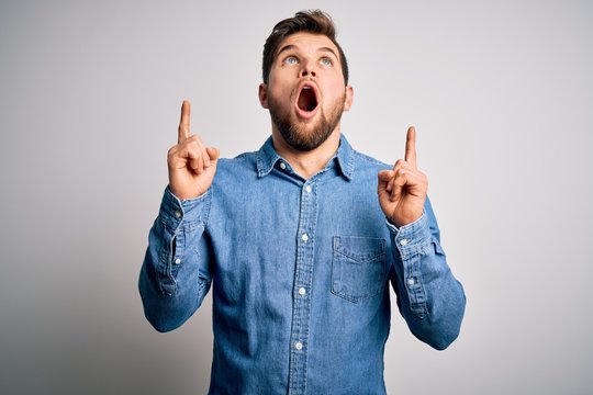 Young handsome blond man with beard and blue eyes wearing casual denim shirt amazed and surprised looking up and pointing with fingers and raised arms.