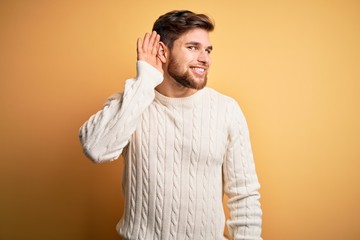 Young blond man with beard and blue eyes wearing white sweater over yellow background smiling with hand over ear listening an hearing to rumor or gossip. Deafness concept.