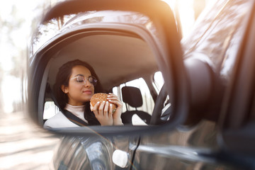 Beautiful brunette model has lunch in a car. Woman eats a hamburger and drinks coffee or tea from a thermos. Food on the road concept.