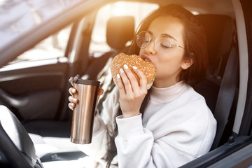 Food on the road concept. Woman eats a hamburger and drinks coffee or tea from a thermos. Food on the road concept.