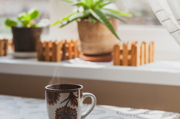 A steaming cup of hot tea or coffee in the early morning on a table near the window. Plants on the windowsill