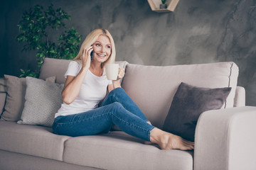 Obraz premium Portrait of her she nice attractive charming cheerful cheery mature lady sitting on divan resting drinking cacao talking on phone in modern industrial loft interior style living-room apartment