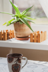 A steaming cup of hot tea or coffee in the early morning on a table near the window. Plants on the windowsill