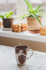 A steaming cup of hot tea or coffee in the early morning on a table near the window. Plants on the windowsill