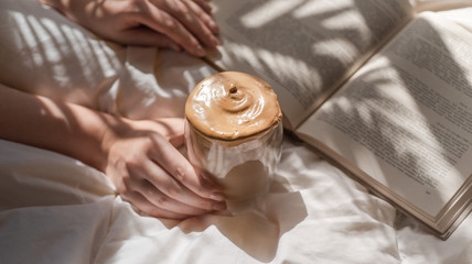Woman hands holding a glass of dalgona coffee and a book in bed with sun glare and palm shadows 