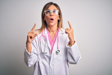 Middle age beautiful doctor woman wearing pink stethoscope over isolated white background amazed and surprised looking up and pointing with fingers and raised arms.
