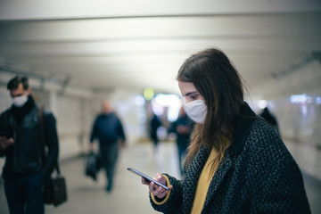 young woman in a protective mask reading a text message at a metro station.
