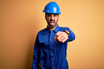 Mechanic man with beard wearing blue uniform and safety helmet over yellow background pointing...